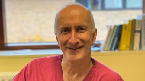 A man in red scrubs smiles into camera, he has white hair on the sides of his head