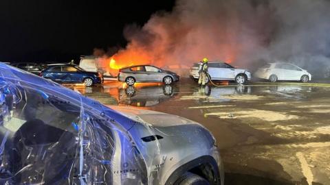 Cars at a salvage yard - some on fire. A firefighter with a long hose is spraying a car with water.