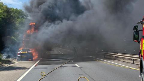 Flames engulf a car transporter which is in the hard shoulder of a dual carriageway. Black smoke billows across the road. A red and yellow fire engine is partially visible on the right of the frame.