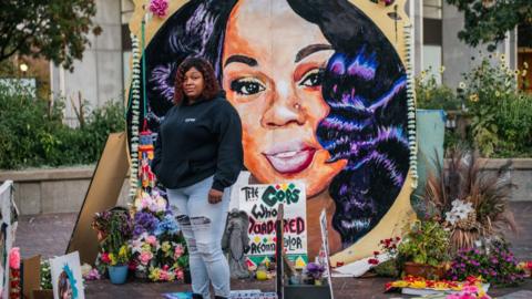 Breonna Taylor's mother stands in front of a mural with her image
