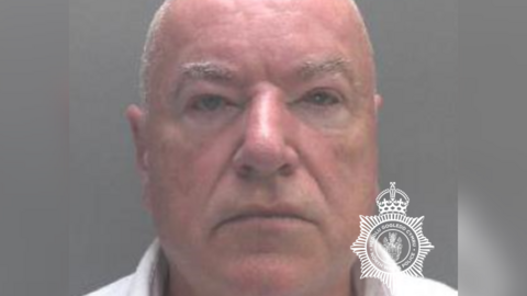 A police mugshot of Neil Foden. He has short white hair and is looking directly into the camera. He is almost entirely bald, with fine, grey very cropped hair on the sides. The background is all grey and he is wearing a white collared shirt