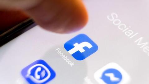 A thumb hovers over a touchscreen which shows applications for Facebook, a blue square with a white F, and Facebook messenger, a white square with a blue speech bubble and a white horizontal zig-zag in the middle. The folder for the apps reads "Social Media" at the top of the screen. A third app below shows a ringing phone in a speech bubble in a blue square.