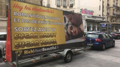 A large billboard in French shows a couple apparently having sex in a high-end modelled photograph, accompanied by French text which reads: "Hey students! Romance, passion, and no student loan, meet a Sugar Daddy or a Sugar Mama"