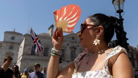 A woman stands in central London. She is wearing a strappy dress, with her hair in a high ponytail with a plait, large white flowery earrings and sunglasses on shields her face from the sun with an orange and white patterned fan. 