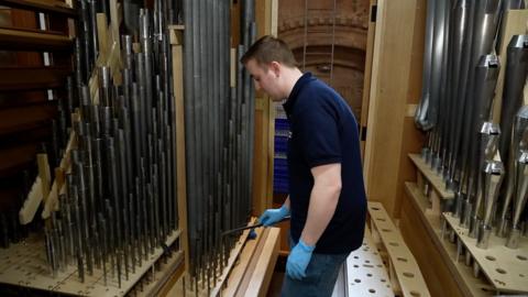 A man holding a tool tunes one of a row of hundreds of organ pipes.