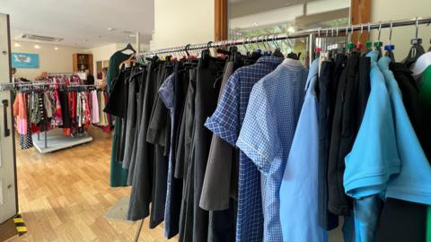 Interior of a clothing store with wooden flooring, featuring racks of shirts and jackets in the foreground, including blue checkered and solid-colored tops. Additional racks with dresses and other garments are visible in the background. The store is well-lit with natural light.