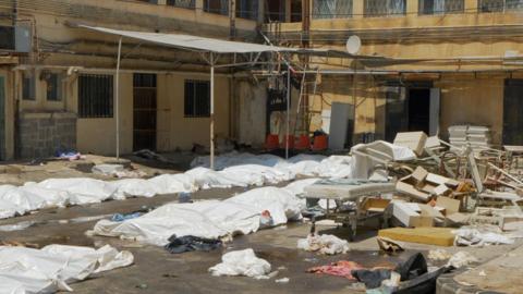 White plastic sheets cover bodies in the carpark of a hospital in Suweida. There is debris of chairs and hospital beds scattered across the ground too