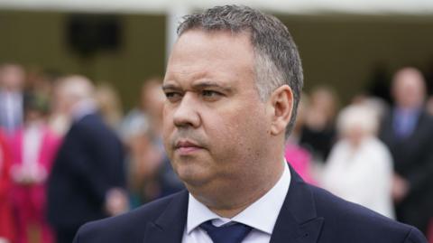 John Caldwell is standing in front of a group of people. He is wearing a navy suit with a white shirt