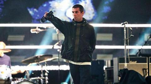 Liam Gallagher pointing with his right hand into the crowd at an Oasis concert. He is holding a tambourine in his right hand and is wearing a dark-coloured jacket.
