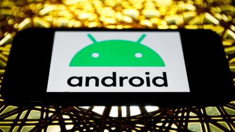 The Android logo on a phone screen in landscape mode, placed on an elaborate metal table lit in golden light