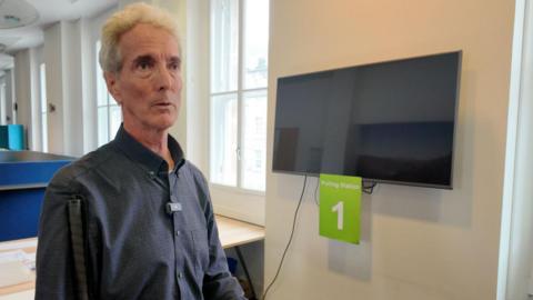 Ken Reid, a mature man with short white hair and a navy checked shirt stands in front of the mock-up polling station Edinburgh City Council has created for training. It is a bright room with windows along one wall.