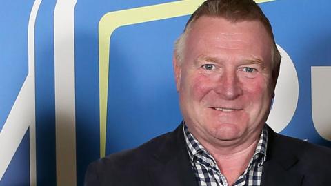 Jim Gracey looking at the camera and smiling. He has short hair and is wearing a black and white checked shirt and a dark blazer