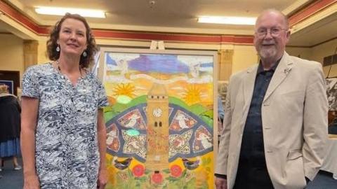 A woman and a man are stood either side of a painting which depcits a town's war memorial. The painting is on an easel, in a room with other artwork being displayed.