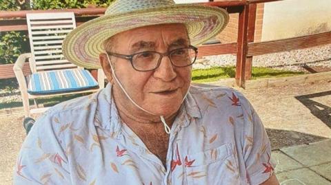 Donald Burgess wears a straw sun hat tied under his chin and a white shirt with yellow and red petal-type shapes on it. He is sitting outside on a patio and is wearing glasses.