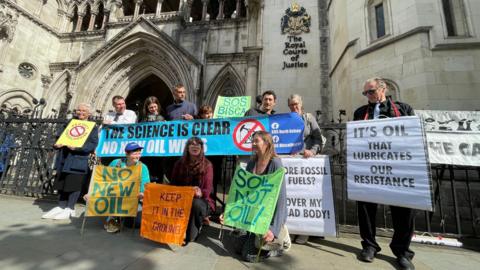 Several campaigners from SOS Biscathorpe pose with colourful placards outside the Royal Court of Justice. Words on some of the placards read "it's oil that lubricates our resistance," "keep it in the ground", "no new oil".
