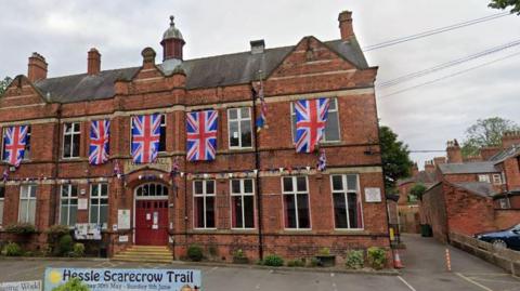 A Google screenshot view of Hessle Town Hall. It shows a red-brick town hall decorated with large Union flags and bunting. A banner in front reads "Hessle Scarecrow Trail". It has large windows and a large red door. There are parking spaces in front of the building. The sky is blue and cloudy.