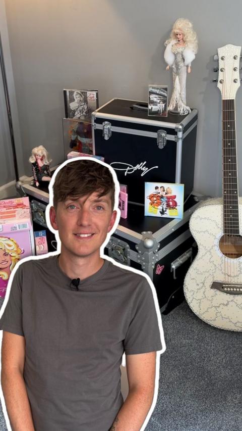 A man with brown hair wearing a grey shirt outlined in white in front of Dolly Parton memorabilia