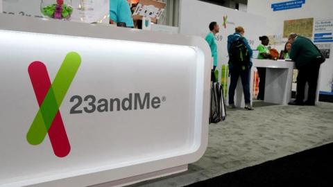 The 23andMe logo is seen at a genealogical convention. The logo is on a bright, white display stand at the front of a carpeted exhibition stall for the company, with people in light blue uniform shirts and others in plain clothes around a table in the background.