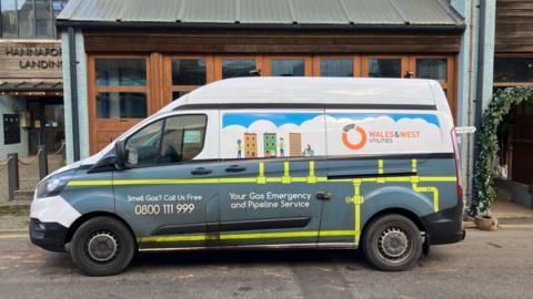 The side of a Wales and West Utilities van. The vehicle is parked on the side of a road, in front of businesses. The van has the gas utility logo printed on its side. It says 'your gas emergency and pipeline service'.
