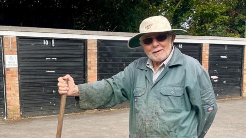An elderly man with a white beard, dark sunglasses, a white cricket hat and green overalls covered in paint blotches is standing in front of five garages. He is holding a broom in one hand and trees can be seen in the background.