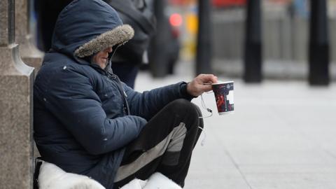 A man wearing a blue parka jacket and black jogging bottoms sits on a duvet on the street while holding out an empty coffee cup to passers by