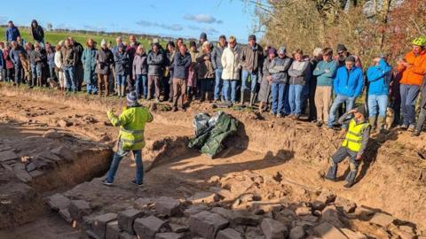 Crowds line the outside of a trench containing several blocks of stone from Hadrian's Wall. A man in a hi-vis yellow jacket is standing in the middle and talking to them.