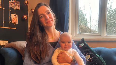 Laura Fenton is sat on a blue sofa, with daughter Alana in her arms.
