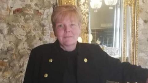 Head and shoulder shot of a woman with blonde hair and wearing a black coat looking straight at the camera. A mirror with a gilded border can be seen to her left