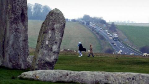 People walking past Stonehenge as cars drive down the A303 in the distance
