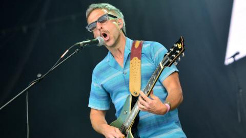  Irwin Sparkes of The Hoosiers performs on stage at Penn Fest on July 23, 2022 in Penn Street, Buckinghamshire. He is wearing sunglasses, a blue short sleeve shirt and holding a guitar as he sings into a microphone.