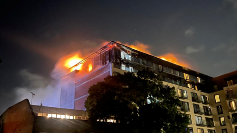 The Mosaic Apartment on fire . The top apartment of a block of flats has flames coming from the roof. It is night. In the foreground you can see a tree and a second brick building.