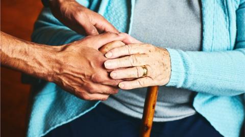 A younger person placing their hands onto the hands of an older person, which are sitting on a cane