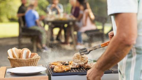 Generic image of a man cooking meat on a barbecue with his family seated at a table in the background