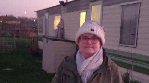 Jaimee Bremer wearing a hat, scarf and coat, stands outside her caravan in the early morning