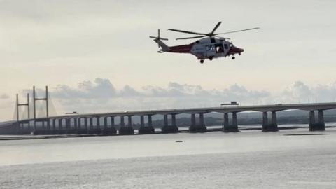 A helicopter hovering over the Severn Estuary with the Prince of Wales Bridge in the background