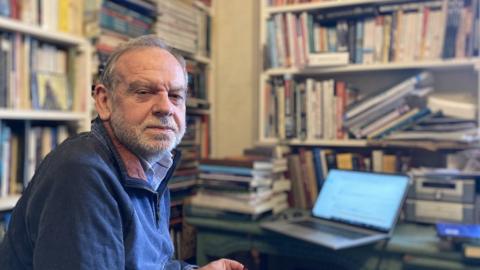 Tony McAleavy looks into the camera, sitting in front of his laptop and shelves stacked with history books