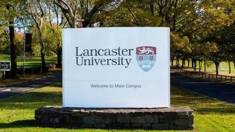 Lancaster University sign in the grounds of the university. It can be seen on a strip of green grass, with two sets of trees on either side. 