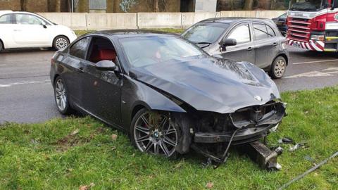 A car which has been damaged in a crash. It is sitting by the side of a road on a grass verge.