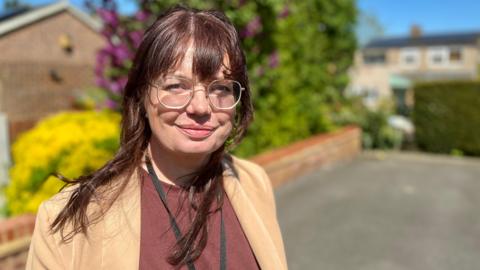 Claire Colvill, a social work apprentice, looks at the camera outside a client's home in Hertfordshire. She is wearing a light brown suit jacket with a maroon smart T-shirt underneath it. She has a black lanyard around her neck and is wearing metal framed glasses. She has brown hair with a fringe that has a red tone. She is standing in front of a driveway with a brick wall and hedges along the wall.