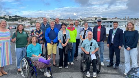 Picture of organisers, deputies and civil servants who took part in the event. Two people are wheelchair users. The rest of the group are stood in a row behind the wheelchair users. They are all smiling at the camera. 