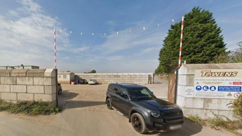 A black car is leaving a car park on an industrial estate next to a sign saying Towens. Concrete block walls surround the site.