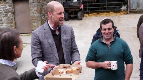 The Prince of Wales wearing a grey suit jacket stood in a courtyard of a farm. He is holding a cardboard box of baked goods. A lady in a grey fleece coat laughs to his left and a man in a green quarter zip jumper holding a cup of tea smiles and laughs to his right.