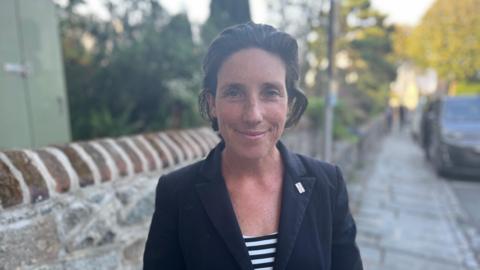 Deputy Lindsay de Sausmarez - A woman with short brunette hair, looking at the camera. She's wearing a black jacket with a sideways Guernsey flag lapel pin on her left lapel. She is also wearing a low black and white striped blouse.