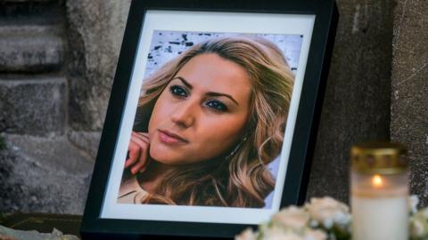 A photo frame showing a picture of Viktoria Marinova is seen laid against the stone walls of a church, with candles in tribute placed nearby