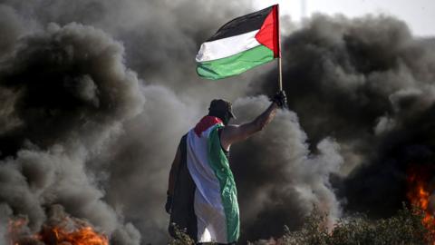 A Palestinian protester waves a Palestinian flag while standing near black smoke billowing from burning objects during a protest near the Gaza-Israel perimeter fence (22 September 2023)