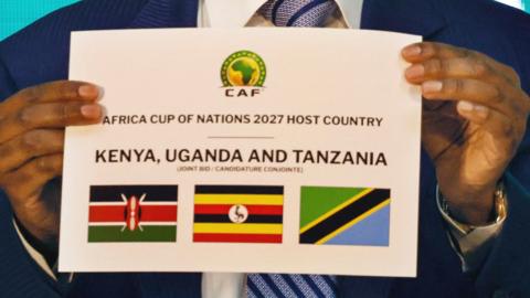 Patrice Motsepe holds up a card with flags of Kenya, Uganda and Tanzania as he announces host of Afcon 2027