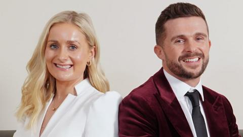 A man in a red velvet jacket on the the right sits with his back to a blonde woman in an all-white pantsuit. Both are smiling and look very smartly well turned-out in a photoshoot/studio setting