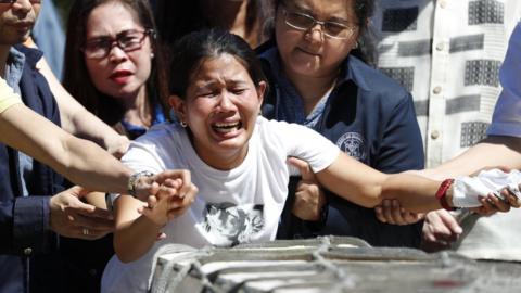 A distraught woman, held by those around her, grieves in front of a long wooden box carrying her sister's remains