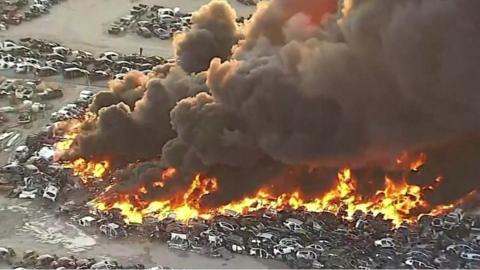 Cars at a junkyard on fire sending a thick cloud of smoke into the air