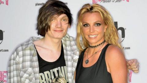 Kurtis standing with Britney, with his arm behind her. Kurtis is wearing a checked white, grey and green shirt, open buttoned, with a top underneath which has Britney spelt in green and an animated image of her on the top. Britney is smiling wearing a black vest top and white trousers, with a black neck choker and a necklace. The background is of a white advert board with promotional branding.